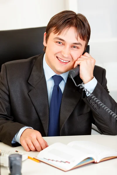 笑顔の近代的な実業家のオフィスの机に座っていると電話で話しています。 — ストック写真
