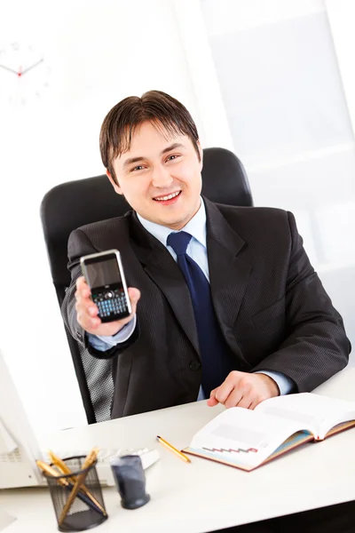 Χαμογελώντας επιχειρηματία που κάθεται στο γραφείο και κρατώντας το κινητό τηλέφωνο με το β — Φωτογραφία Αρχείου