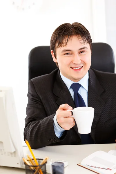 Χαμογελώντας επιχειρηματία που κάθεται στο γραφείο και κρατώντας το φλιτζάνι τσάι στο χέρι — Φωτογραφία Αρχείου