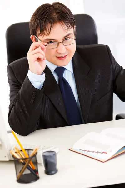 Empresário sorridente sentado na mesa do escritório e endireitando óculos — Fotografia de Stock