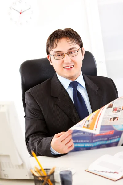 Hombre de negocios sonriente sentado en el escritorio de la oficina y sosteniendo el periódico en las manos — Foto de Stock