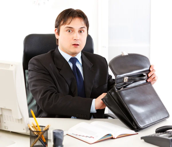 Shocked modern businessman sitting at office desk and holding open suitcase — Stock Photo, Image