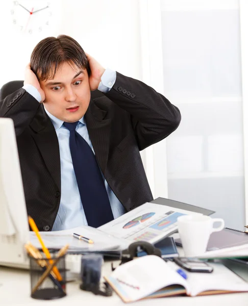 Shocked young businessman sitting at office desk being overloaded with loa — Stock Photo, Image