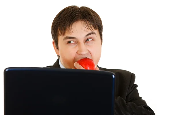 Homem de negócios moderno cautelosamente olhando para fora do laptop e comer maçã — Fotografia de Stock