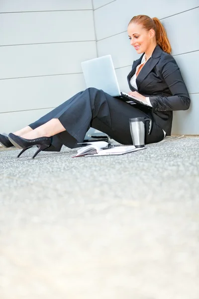 Smiling business woman sitting on floor at office building and using lapto