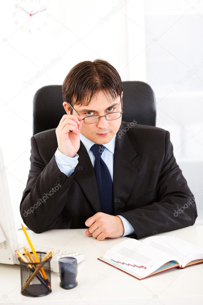 Authoritative businessman sitting at office desk and straightening eyeglas