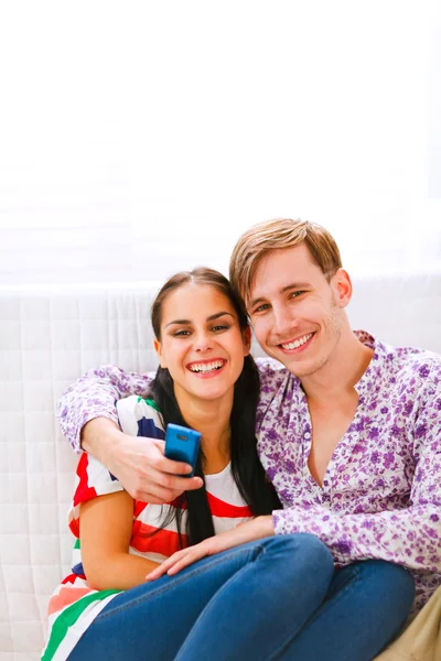 Jovem casal sorrindo sentado no sofá com celular — Fotografia de Stock