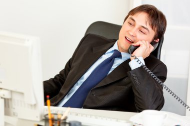 Pleased modern businessman sitting at office desk and talking on phone clipart