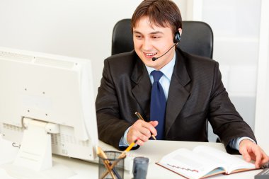 Smiling businessman with headset sitting at office desk and looking at co clipart