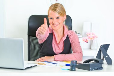 Smiling business woman sitting at office desk and showing thumbs up clipart