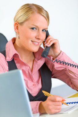 Smiling business woman sitting at office desk and talking on phone clipart