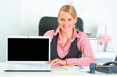 Smiling business woman sitting at office desk and showing laptop with blank clipart