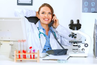 Relaxed doctor woman sitting at office table and talking on phon clipart