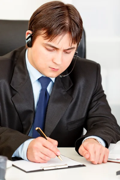 Concentrated businessman with headset sitting at office desk and taking n — Stock Photo, Image