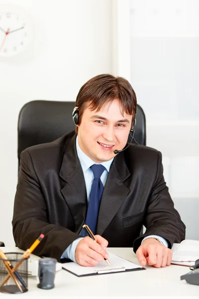 Lächelnder Geschäftsmann mit Headset sitzt am Schreibtisch und macht sich Notizen — Stockfoto