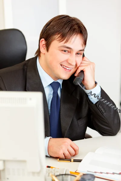 Sonriente hombre de negocios moderno sentado en el escritorio de la oficina y hablando por teléfono — Foto de Stock