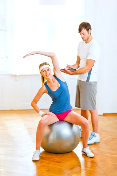 Chica sonriente haciendo ejercicios en la pelota de fitness debajo de la supervis —  Fotos de Stock