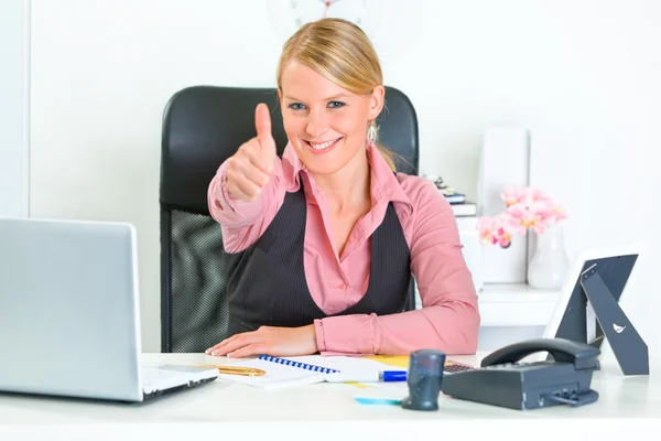 Sorrindo mulher de negócios sentado na mesa do escritório e mostrando os polegares para cima — Fotografia de Stock