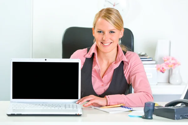 Улыбающаяся деловая женщина сидит за рабочим столом и показывает ноутбук с пустым — стоковое фото