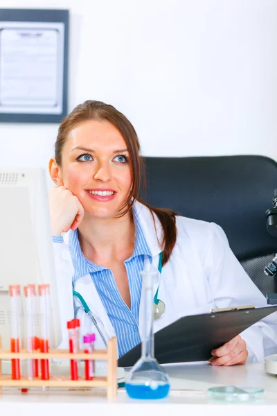 Mujer doctora sonriente sentada en la mesa del consultorio y mirando en la esquina — Foto de Stock