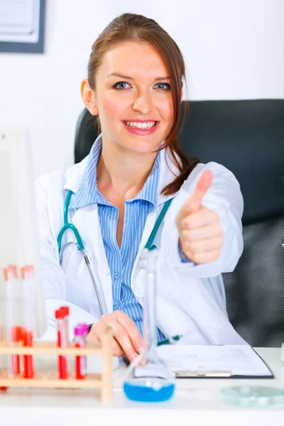 Mujer doctora sonriente sentada en la mesa del consultorio y mostrando el gesto de los pulgares hacia arriba —  Fotos de Stock