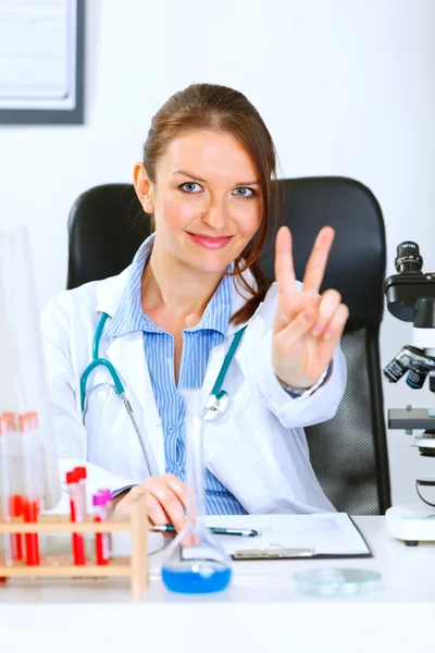 Mujer doctora sonriente sentada en la mesa de la oficina y mostrando la victoria — Foto de Stock