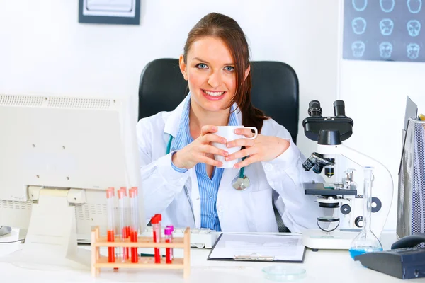 Médico sonriente mujer sentada en la mesa de la oficina y sosteniendo la taza — Foto de Stock
