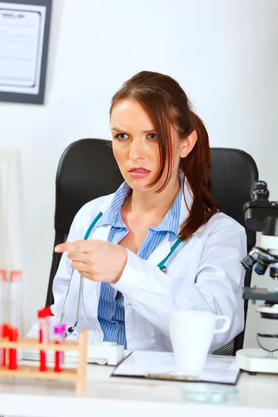 Запутавшаяся женщина-врач сидит за рабочим столом и указывает пальцем — стоковое фото