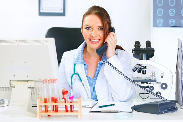 Mujer doctora sonriente sentada en la mesa de la oficina y hablando por teléfono —  Fotos de Stock