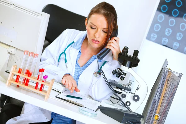 Concerned doctor woman talking on phone and looking in clipboard — Stockfoto