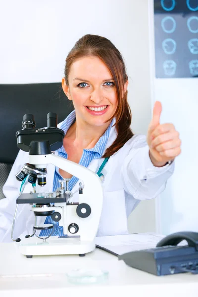 Usmívající se žena lékař pomocí mikroskopu v laboratoři a ukazuje palec nahoru g — Stock fotografie