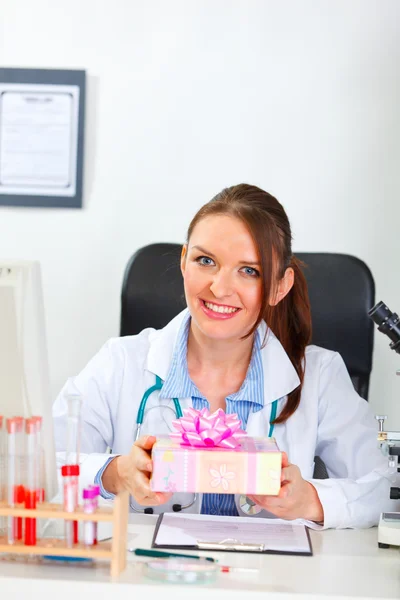 Sorridente medico femminile seduto in ufficio e in possesso di presente — Foto Stock