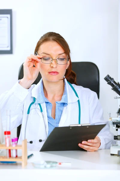Doctor woman sitting at office table and looking in clipboard — Stock Photo, Image