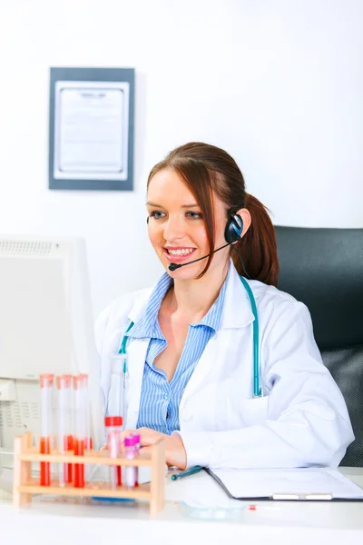 Leende läkare kvinna med headsetet sitter vid office bord och arbetar på co — Stockfoto