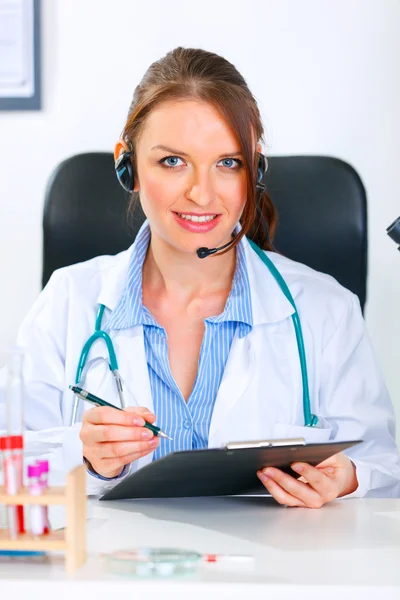 Femme médecin souriante avec casque assis à la table du bureau et tenant clip — Photo