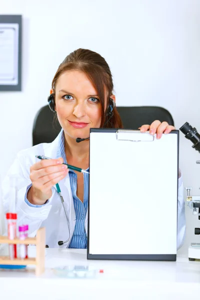 Doctor woman sitting at office table showing blank clipboard and — Stock Photo, Image