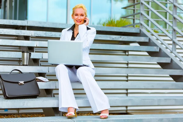 Smiling business woman sitting on stairs with laptop and talking — Stock Photo, Image