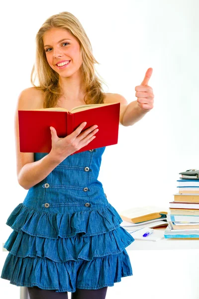 Sonriente chica con el libro en la mano de pie cerca de la mesa y mostrando los pulgares hacia arriba ge — Foto de Stock