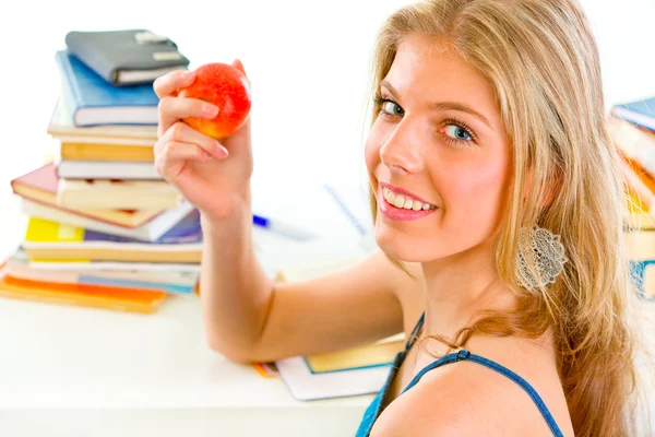Sonriente chica adolescente sentada en el escritorio con libros y sosteniendo la manzana en la mano — Foto de Stock