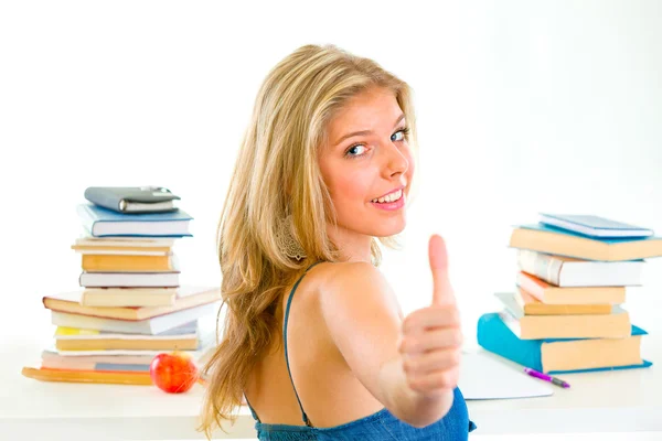 Lachende tiener meisje zit aan bureau met boeken en tonen duimschroef opwaarts gebaar — Stockfoto