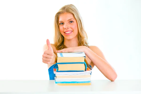 Sorrindo menina adolescente sentada à mesa com livros e mostrando polegares para cima gestur — Fotografia de Stock
