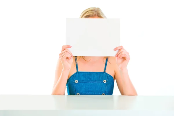 Девушка сидит за столом и держит пустую бумагу перед лицом — стоковое фото