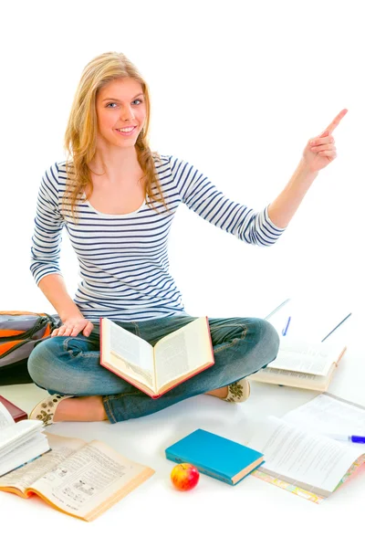 Улыбающаяся девочка-подросток, сидящая на полу среди учебников и указывающая — стоковое фото