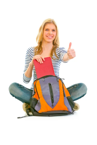 坐在地上微笑的女孩从书包中获取的书和显示的拇指 — 图库照片