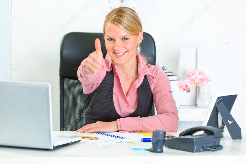 Smiling business woman sitting at office desk and showing thumbs up