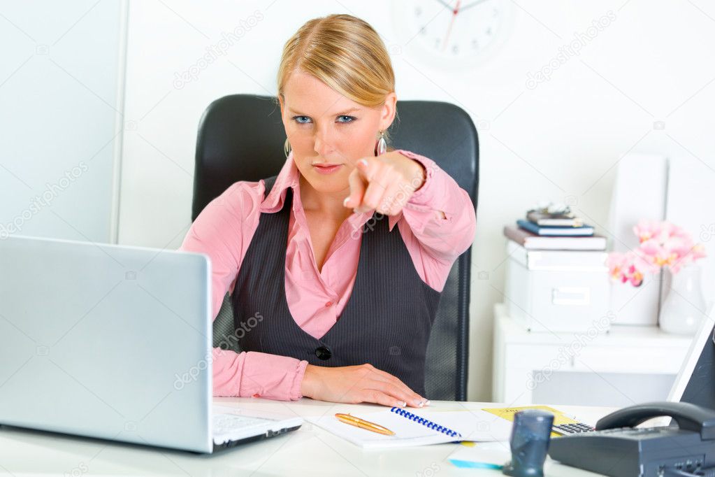 Confident business woman sitting at office desk and pointing finger at you