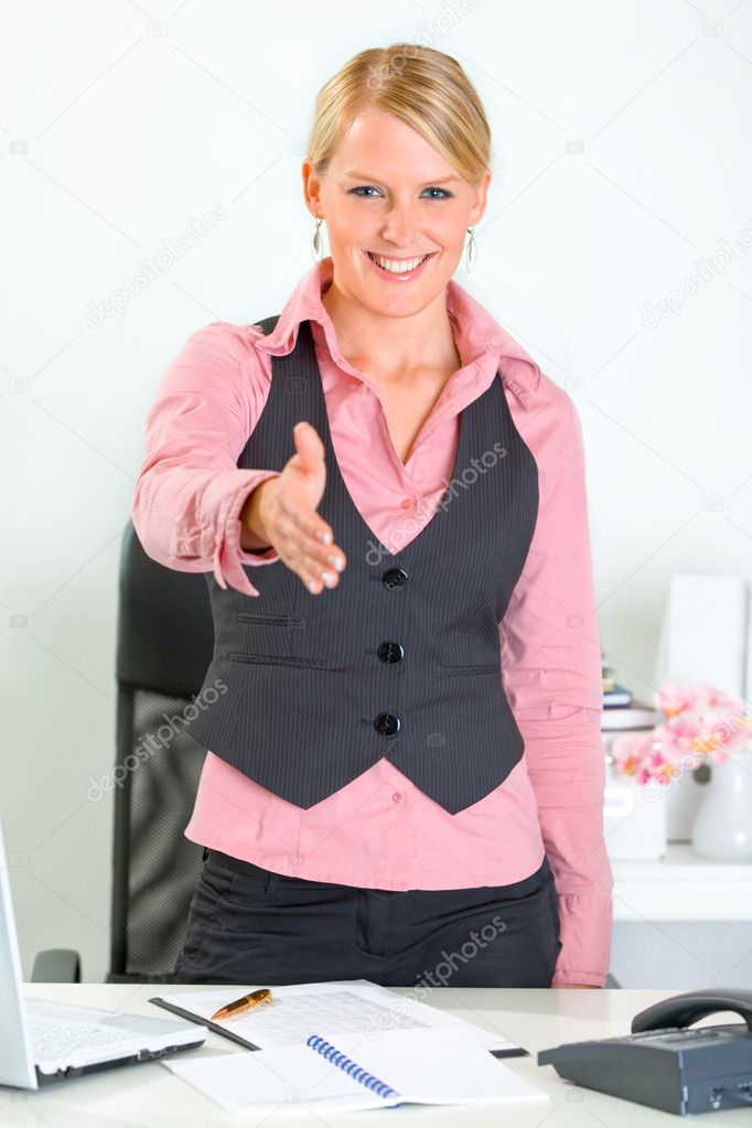 Smiling business woman standing near office desk and stretches out hand for