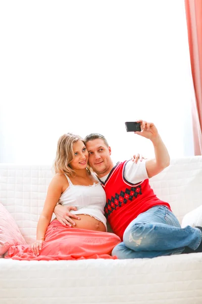 Hermosa embarazada sentada con el marido en el sofá y tomando autorretrato —  Fotos de Stock