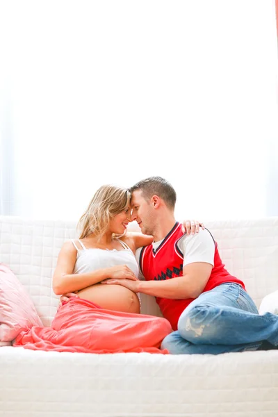 Счастливая беременная женщина сидит с мужем на диване и обнимается — стоковое фото