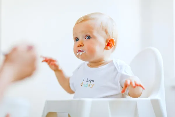 Eat smeared lovely baby in baby chair feeding by mother — Stock Photo, Image